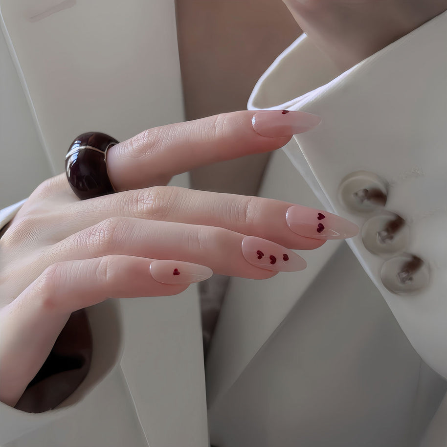 Glossy medium almond nails with nude base and tiny red hearts for a soft, romantic vibe.