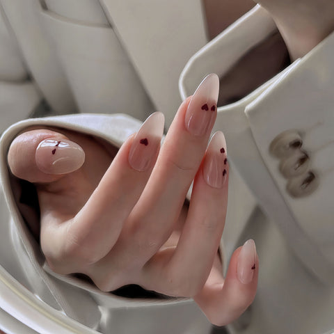 Glossy medium almond nails with nude base and tiny red hearts for a soft, romantic vibe.