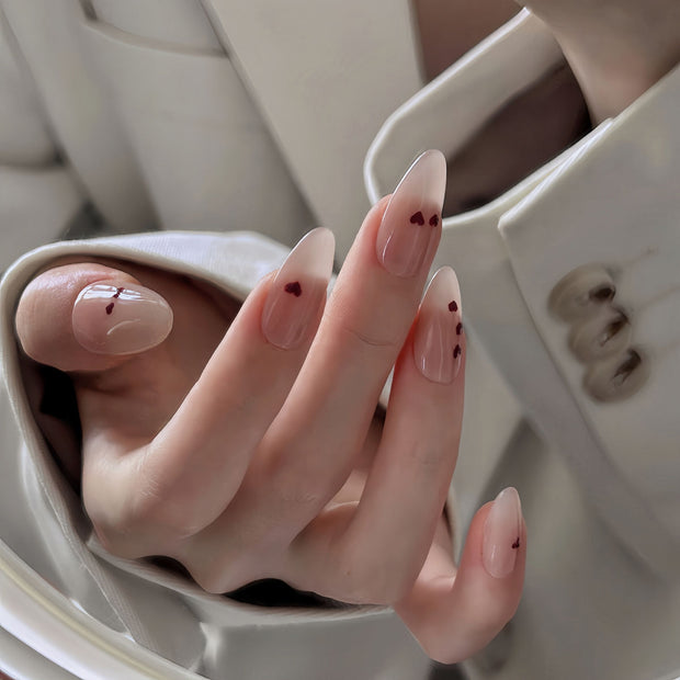 Glossy medium almond nails with nude base and tiny red hearts for a soft, romantic vibe.