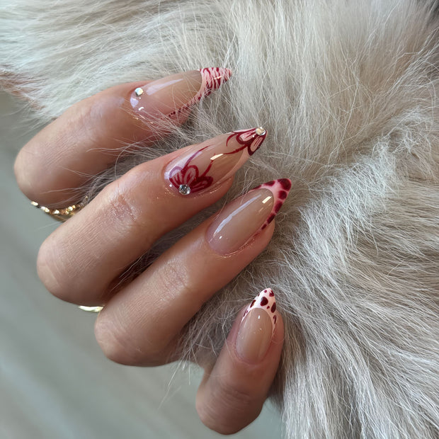 Glossy medium almond nails with nude base, red swirl patterns, and crystal details for a romantic, edgy vibe.