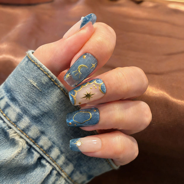Celestial-inspired press-on nails: nude-blue gradient with gold star/moon motifs, perfect for parties.